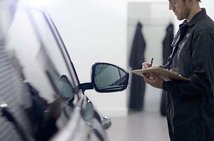 Auto technician in a garage inspecting a black car and taking notes on a clipboard.