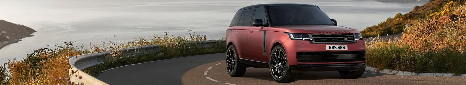 Red Range Rover on a coastal road with scenic ocean and hills in the background, plate RBS 689.